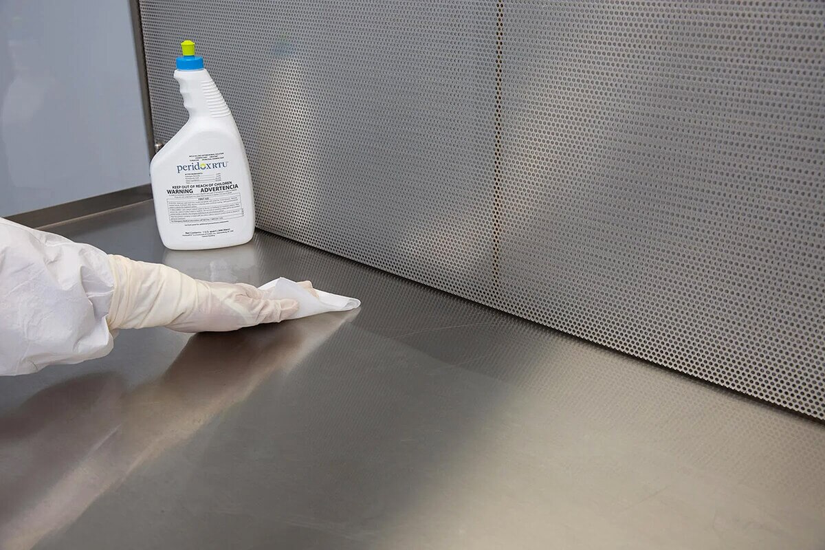 Technician wiping stainless-steel work surface using Peridox RTU sporicidal disinfectant during aseptic cleanroom cleaning procedure.