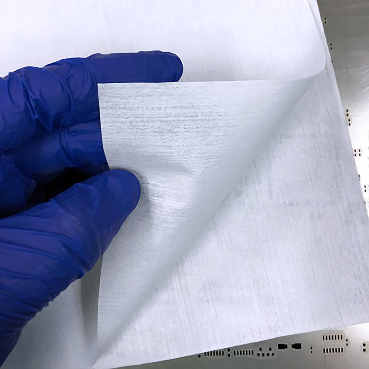 Gloved hand lifting single AstroWipe wipe showing thin white poly-cellulose texture