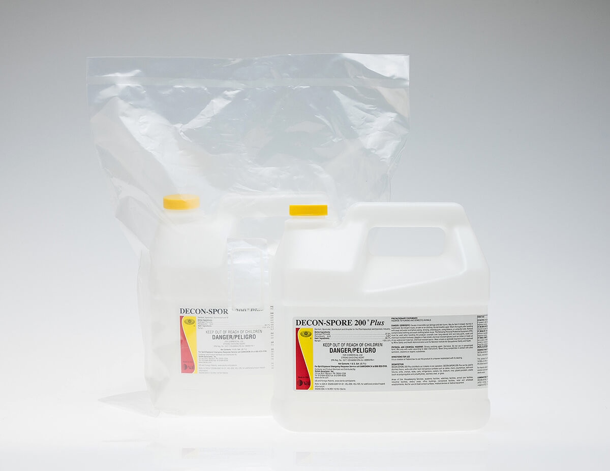 Two DECON-SPORE 200 Plus 1-gallon concentrate containers, one sealed in a clear poly bag and one unbagged in front, displayed on a white reflective surface.