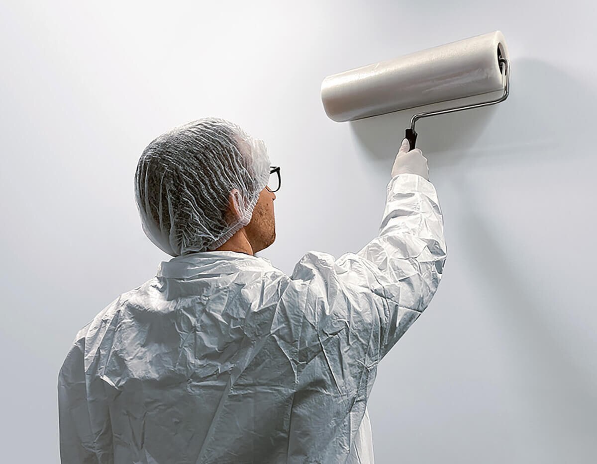 Person in cleanroom attire using the 18" Tacky Traxx roller on a wall, demonstrating how the foam refill attaches to a roller frame and removes particles from textured surfaces.